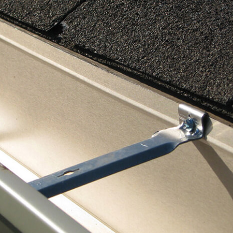 Close-up of a blue metal support brace bolted to a beige wall under dark roof shingles.