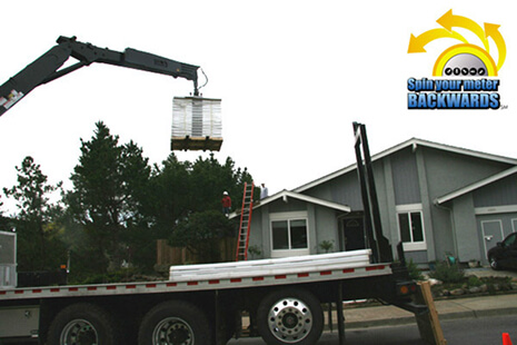 Flatbed tow truck with a crane lifting a pallet toward a suburban house during a delivery/construction.