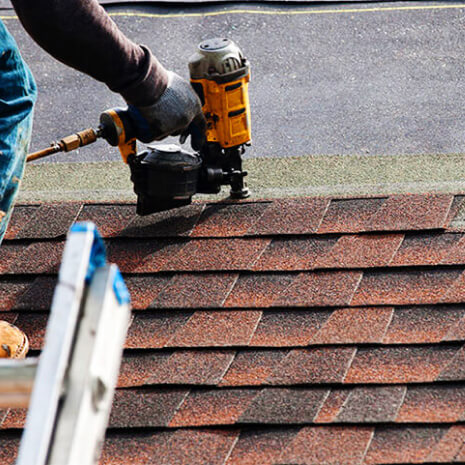 Worker wearing a glove uses a cordless nail gun to fasten brown roof shingles; a ladder rests nearby.