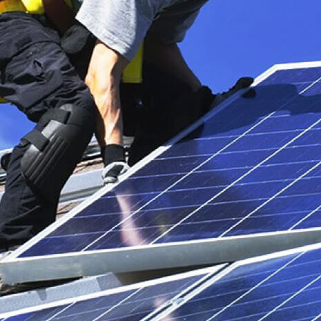 Worker in safety gear installing blue solar panels on a rooftop under a clear sky