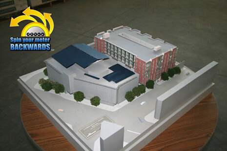 3D architectural model of a building complex on a round pedestal, with white and brick structures and trees; includes a 'Spin your meter backwards' logo in the top-left.