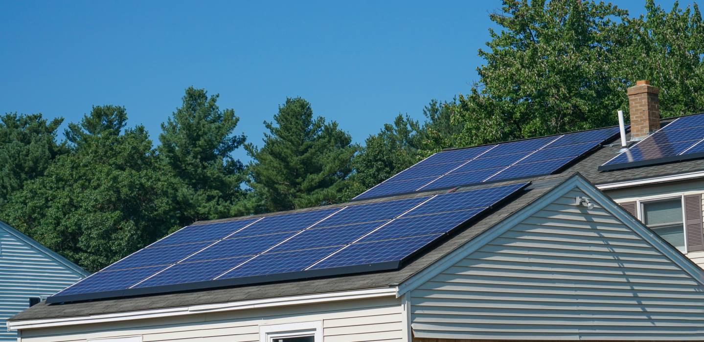 Residential house with blue solar panels on the roof, blue sky and green trees in the background.